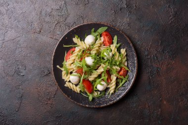 Çeri domatesli lezzetli vejetaryen makarna, yumuşak peynirli mozzarella ve koyu beton arkaplanda yeşil roka yaprakları.