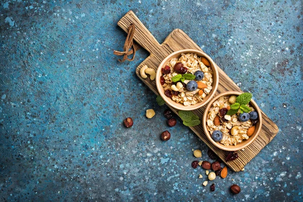新鮮なブルーベリー アーモンド ヘーゼルナッツとカシューナッツとオートミールのトップビュー 木製ボード上の健康的な朝食のための緑のミントの葉と青の具体的な背景 — ストック写真