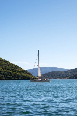 Yelkenli, berrak bir gökyüzünün altındaki yeşil tepeler arasında durgun mavi sularda sürükleniyor. Huzurlu deniz manzarası, manzaralı bir kıyı manzarasında özgürlük, seyahat ve rahatlık yakalıyor..