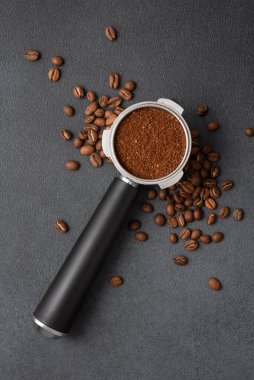 Portafilter koyu dokulu bir yüzey üzerinde kavrulmuş fasulyelerle çevrili öğütülmüş kahveyle dolu. Hayat yoğunluğu, enerjiyi, işçiliği ve odaklanmış espresso hazırlama ritüelini vurgular..