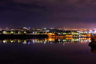 Mor gökyüzü ve şehir ışıkları. Gece şehrinin parıldayan evleri göle yansıyor. Evleri, gölü ve inanılmaz güzel gökyüzü olan güzel bir şehir manzarası. Violet gökyüzü