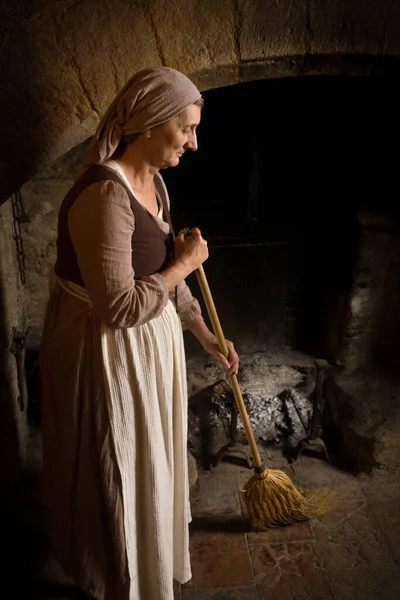 在法国 一名身着文艺复兴女装的妇女用扫帚清扫厨房的壁炉 这间壁炉是一座正宗的中世纪城堡 — 图库照片