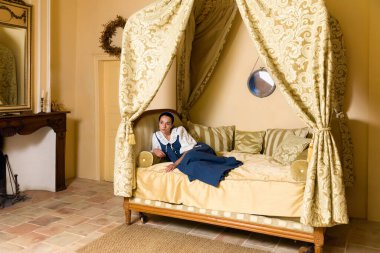 Ortaçağ Fransız şatosunun altın yatak odasındaki lüks bir antika tepe örtüsünün üzerinde duran otantik rönesans kostümü giymiş güzel bir kadının Hollanda Ustası tarzı portresi.