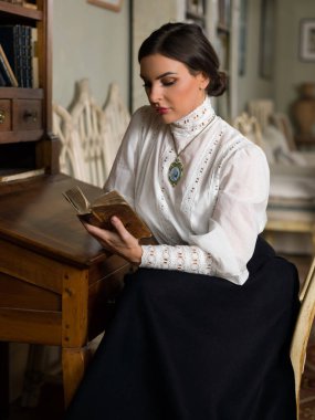 Kütüphanede oturan Edwardian etekli ve yüksek yakalı dantel bluzlu güzel bayan. Öğretmen de olabilir, müdire de..
