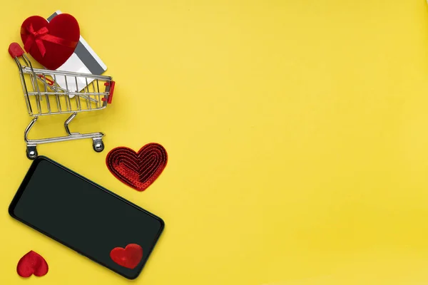 Carro de la compra con caja de regalo, tarjeta de crédito, corazones y teléfono sobre fondo amarillo —  Fotos de Stock