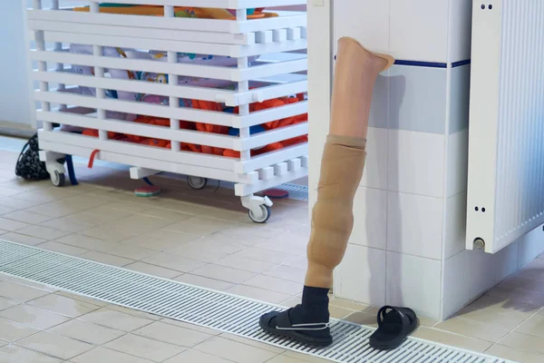 human leg prosthesis stands by the pool wall along with slippers while its owner swims