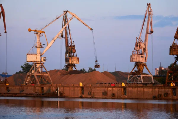level-luffing bulk-handling cranes load sand onto a dry cargo barge in a river port in the evening ligh