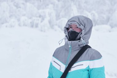young woman in a warm jacket with a hood and a buff neck gaiter on her face against the background of blurred winter landscape clipart