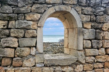 Arkasında denizi görebileceğiniz antik bir duvarın kalıntıları arasında yarı dairesel bir pencere açılıyor.