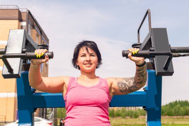 Genç bir kadın göğüs presi makinesiyle dışarıda egzersiz yapıyor. 