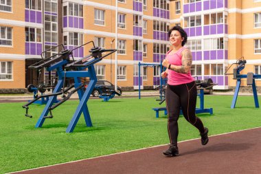 Bir kadın spor sahasında, bir şehir evinin avlusunda sokak spor aletlerinin arka planında koşuyor.