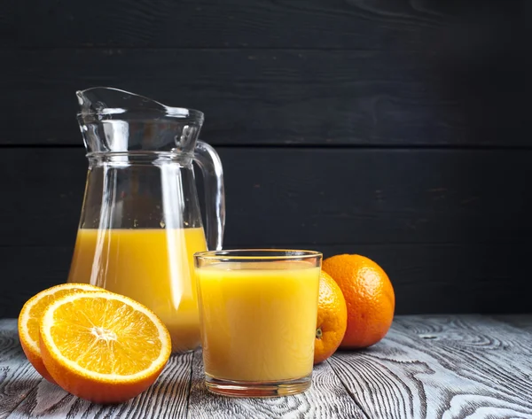 Copo de suco de laranja fresco, jarro e laranjas maduras na mesa de madeira — Fotografia de Stock