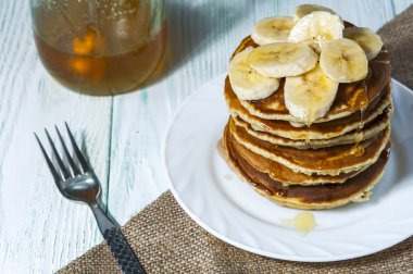 Muz dilimleri ve çatal ve keten peçete ahşap arka plan üzerinde beyaz tabakta bal ile ev yapımı krep yığını