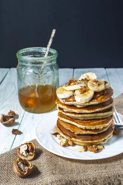 Stack of homemade pancakes with banana slices and honey on white plate with fork and linen napkin on wooden background. — Stock Photo, Image