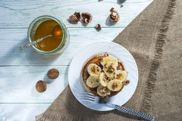 バナナのスライスと木製の背景にフォークとリネンのナプキンで白い皿の上に蜂蜜の自家製パンケーキのスタック. — ストック写真