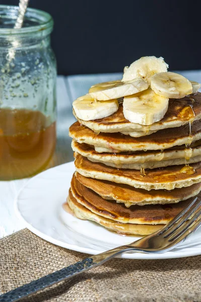 Stapel huisgemaakte pannenkoeken met plakjes banaan en honing op wit bord met vork en linnen servet op houten achtergrond. — Stockfoto