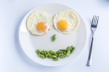 Two fried eggs with green beans on white plate, fork on light background. English breakfast.