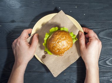 Appetizing hamburger with vegetables on white plate with fork and knife on wooden table. Fast food, junk food. Hamburger in woman's hands. Unhealthy food concept.