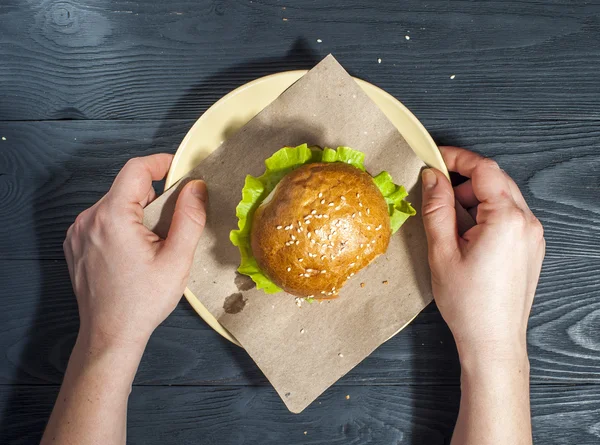 Appetizing hamburger with vegetables on white plate with fork and knife on wooden table. Fast food, junk food. Hamburger in woman's hands. Unhealthy food concept. — Φωτογραφία Αρχείου