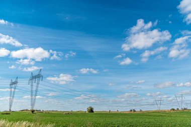 Güneşli bir günde düz ve yeşil bir tarım arazisinde yüksek voltaj hatları ve elektrik direkleri.