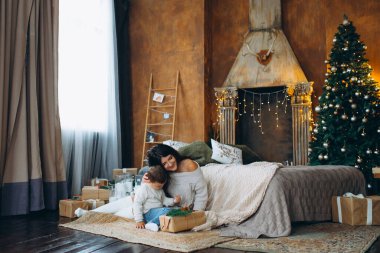 Güzel kahverengi saçlı anne ve sevimli oğlan yerde oturuyor ve Noel ağacının arka planındaki bir kutuda hediyeleri açıyor..