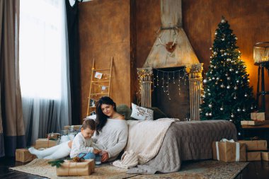 Güzel kahverengi saçlı anne ve sevimli oğlan yerde oturuyor ve Noel ağacının arka planındaki bir kutuda hediyeleri açıyor..