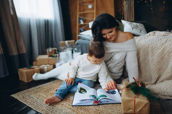 Güzel kahverengi saçlı anne ve sevimli oğlan yerde oturup farklı noel ağaçlarında masal kitapları okuyorlar..