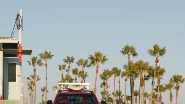 Ikonisk retro-tre-vakttårn og bajonettur-rød bil. Livbøye, amerikansk statsflagg og palmer mot blå himmel. Sommerkalifornia estetikk, Santa Monica strand, Los Angeles, CA USA – stockvideo