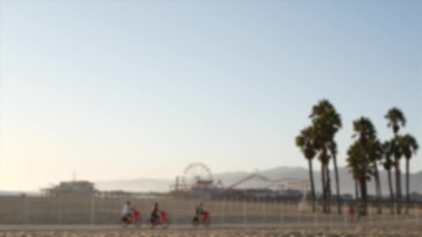 Kalifornische Strandästhetik, die Menschen fahren mit dem Fahrrad auf einem Radweg. Unscharfer, defokussierter Hintergrund. Vergnügungspark auf Pier und Palmen in Santa Monica, USA — Stockvideo