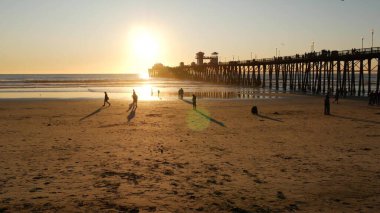 Oceanside, California USA - 27 Aralık 2019: ahşap iskele ve yürüyen insanlar. Turistler deniz kıyısındaki tatil beldesinde geziniyor, yaz tatilleri Los Angeles yakınlarında. Günbatımı okyanus plajı, tropikal güneşli turuncu gökyüzü.