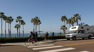 Oceanside, California USA - 8 Şubat 2020: Karavan ve insanlar deniz kenarındaki yolda yürüyor, sörf yapıyor ve sörf yapıyor. Pasifik Okyanusu, tropikal plaj turist beldesi. Sahilde güneşli bir günde, palmiye ağaçları.