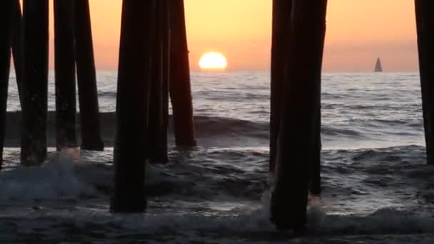 Golven spatten onder de pier, zonsondergang in Oceanside, Californië, USA. Zeewater, zon en houten palen. — Stockvideo