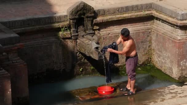 Bhaktapur Nepal October 2018寺院の壁の近くで洗濯する男 通りのレンガの壁に高齢者の石のクレーンからの水で髪や服を洗う若い男のビュー カトマンズの街並み — ストック動画