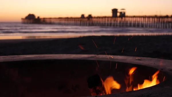 Campfire pit in California USA. Camp fire on twilight ocean beach, bonfire flame by sea water waves.