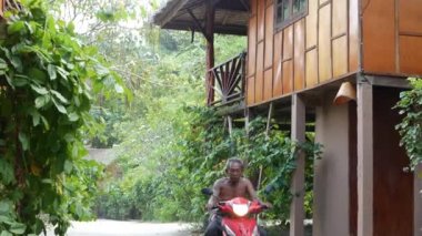 KOH PHANGAN, THAILAND - 11 HAZİRAN 2019 Tropikal ağacın yanındaki Lovely barakalarında motosiklet süren adam. Ormanda güneşli bir günde kurulmuş harika küçük bir köy. Yeşil hayat. Egzotik bir köy. Doğada yaşamak.