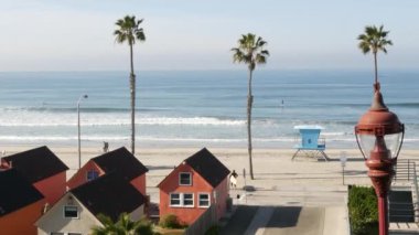 Oceanside, California USA - 20 Şubat 2020: Okyanus dalgalarında sörf yapan sörfçü adam. Renkli sahil kulübeleri, cankurtaran kulesi, gözetleme kulesi. Deniz kenarındaki çok renkli bungalov kulübeleri. Palmiye ağaçları.