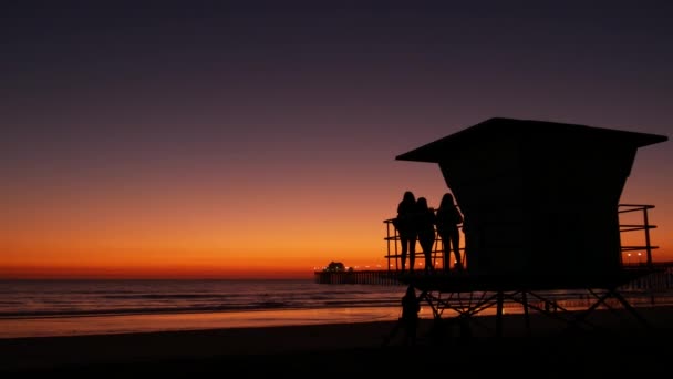 Jovens meninas adolescentes silhuetas, salva-vidas relógio torre, amigos na praia do oceano pacífico, Califórnia EUA. — Vídeo de Stock