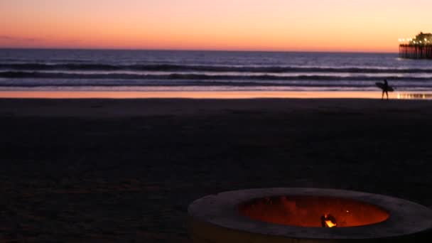 Hoyo de fogata en California USA. Fuego de campamento en la playa del océano crepuscular, llama de hoguera, surfista y olas. — Vídeo de stock