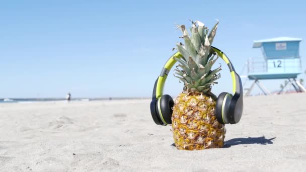 Pineapple in headphones, sandy ocean beach coast. Tropical summer exotic fruit. Ananas on shore. — Stock Video