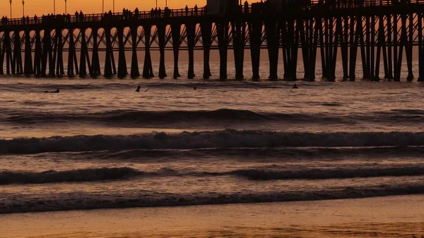 Sylwetka pomostu o zachodzie słońca, Kalifornia, USA, Oceanside. Ośrodek surfingowy, tropikalna plaża oceanu. Surfer czeka na falę. — Zdjęcie stockowe