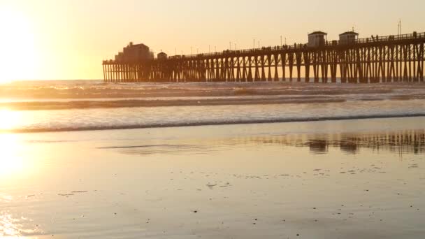 Muelle de madera sobre pilotes, silueta al atardecer, California USA, Oceanside. Olas soleadas al atardecer. — Vídeo de stock