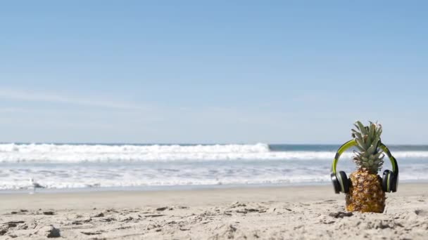 Pineapple in headphones, sandy ocean beach coast. Tropical summer exotic fruit. Ananas on shore. — Stock Video