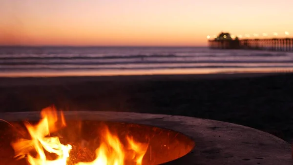 Campfire pit in California USA. Camp fire on twilight ocean beach, bonfire flame by sea water waves.