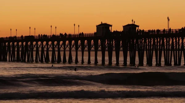 Sylwetka pomostu o zachodzie słońca, Kalifornia, USA, Oceanside. Ośrodek surfingowy, tropikalna plaża oceanu. Surfer czeka na falę. — Zdjęcie stockowe