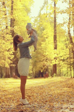 Anne bebeği sonbahar parkına fırlatıyor, mutluluk sarı doğa düşüyor.