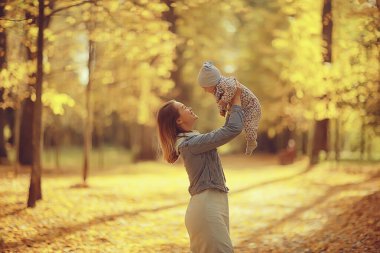 Anne bebeği sonbahar parkına fırlatıyor, mutluluk sarı doğa düşüyor.
