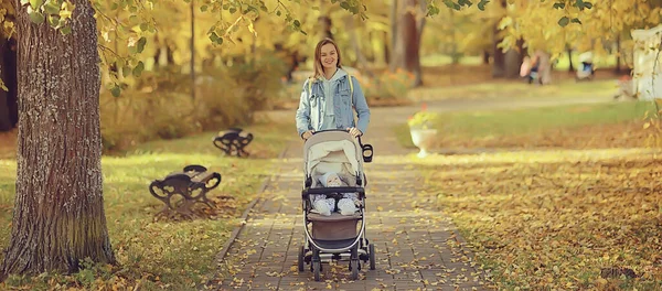 散歩のために秋の公園でベビーカーを持つお母さん風景秋の景色10月の路地黄色の公園 — ストック写真