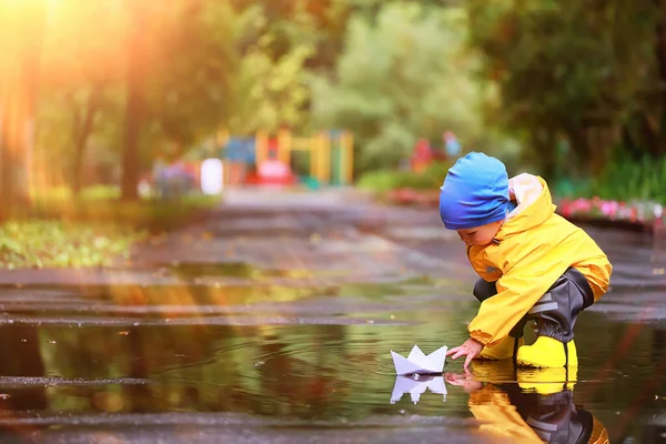 紙のボートの水たまりゲーム 男の子の季節の秋の外観のレインコート 黄色のゴム製のブーツ 屋外ゲーム — ストック写真