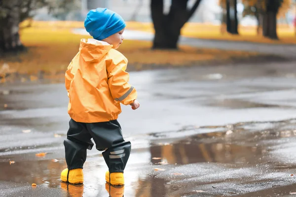 水たまりで屋外で遊ぶ男の子 秋の子供時代のゴム製の靴レインコート黄色 — ストック写真