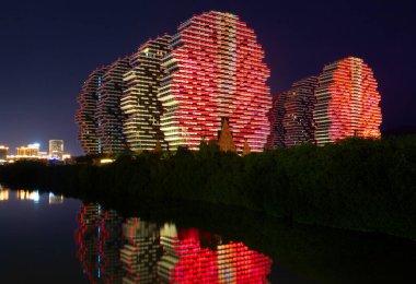 China, Hainan island, Sanya - January 25, 2020: night view from the water to illuminated red lights skyscrapers of the Beauty Crown Grand-Tree Hotel or Houses-Trees. Popular tourism attraction clipart